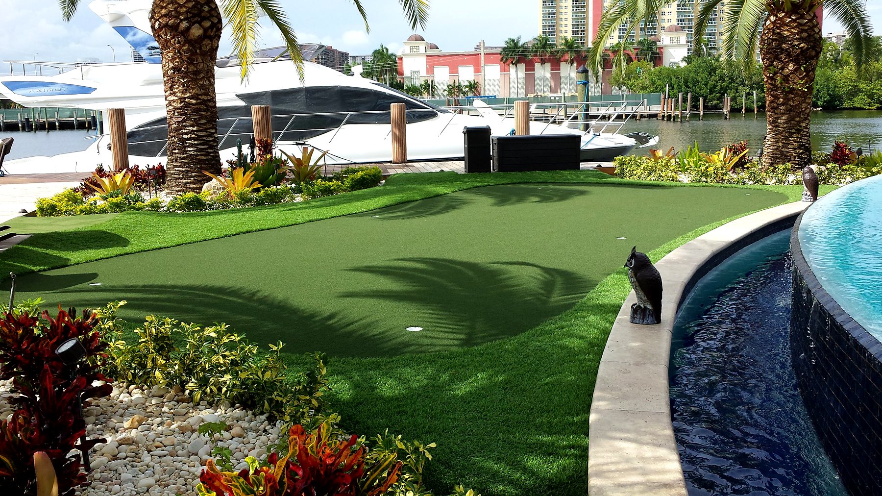 Waterfront backyard putting green with palm trees, a fountain and a docked yacht
